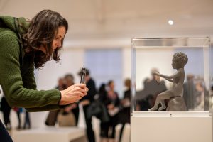 A female figure leaning down and taking a photograph on an iPhone of a cabinet containing a small sculpture by Kiki Smith of a seated female figure holding a flat cloud-shaped object