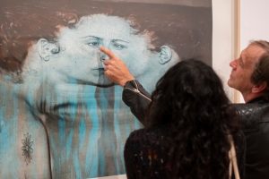 A figure facing and pointing at a print by Kiki Smith depicting her own face stretched left to right in bright turquoise and black