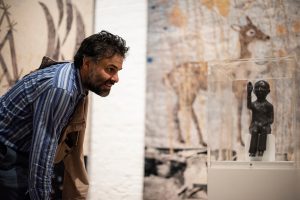 A male figure looking into a cabinet containing a small sculpture of a seated male figure with one arm raised in the exhibition Kiki Smith: I am a Wanderer at Modern Art Oxford