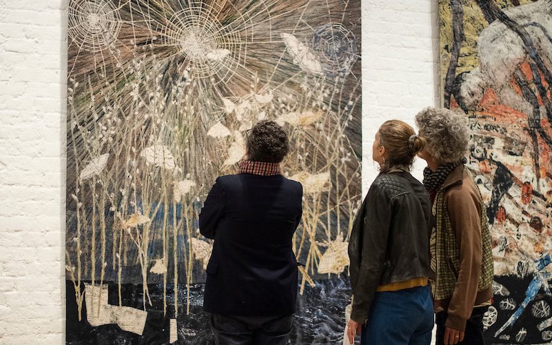Three figures facing a tapestry by Kiki Smith depicting large spider webs and moths in shades of blue and brown in the exhibition I am a Wanderer at Modern Art Oxford