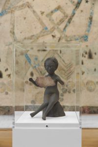 Close up photograph of a small sculpture by Kiki Smith of a female figure sitting on a rock holding an abstract flat cloud-shaped object