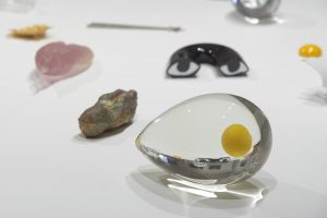 Close up photograph of a glass pointed egg-shaped sculpture with a yellow sphere inside it; and several other small sculptures by Kiki Smith behind