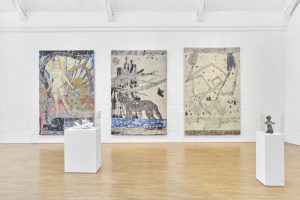 Photograph of three tapestries by Kiki Smith hanging in the Modern Art Oxford exhibition I am a Wanderer. The first depicts a nude female figure surrounded by red and green plants and a snake, the second a blue and black wolf, and the third a geometric pattern featuring owls