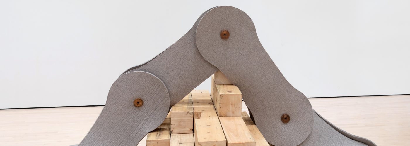 Installation photograph of a large-scale felt sculpture by Johanna Unzueta in the shape of three interlocking cogs that look like parts of industrial machinery. The sculpture forms a an arch over a pile of planks of wood