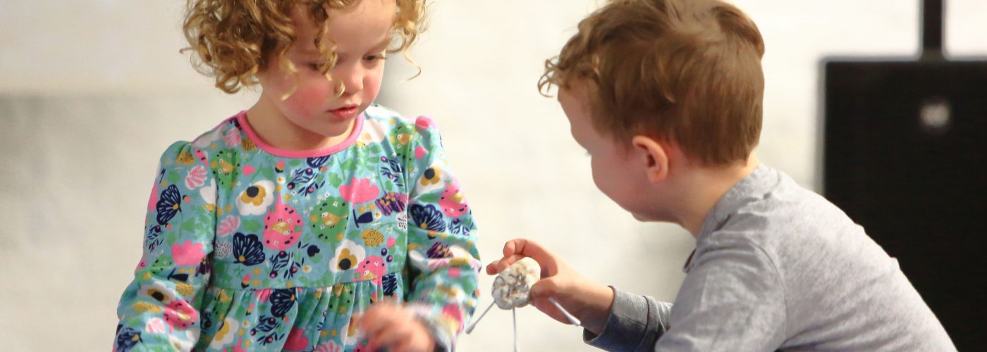 Two children indoors crouching next to a paddling pool. One is showing the other a paper toy