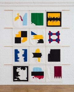 12 brightly coloured squares of fabric hanging on a white brick wall, suspended by wooden sticks. The patterns on the fabric are geometric and reminiscent of maps and flags.