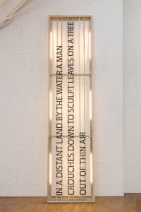 A big wooden frame with large lettering leaning against a white brick wall. There is strip lighting around the edge of the frame and the text reads: In a distant land by the water a man crouches down to sculpt leaves on a tree out of thin air.