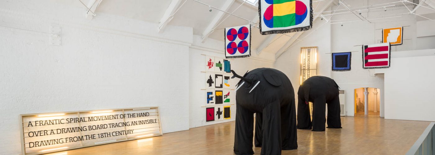 Two large sculptures of elephants surrounded by brightly coloured fabric flags hanging from the ceiling and on the walls of a large industrial space with white walls and a wooden floor.