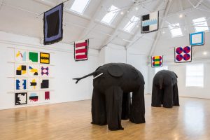 Two large sculptures of elephants surrounded by brightly coloured fabric flags hanging from the ceiling and on the walls of a large industrial space with white walls and a wooden floor.