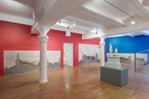 The inside of an art gallery with white columns and a low ceiling. One wall is painted red and the other blue and there are large black and white photographs pasted to the walls.