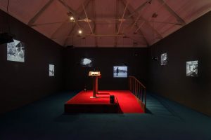 A darkened room in an ex-industrial space. In the centre is a red carpeted raised platform with two lecterns on it, and black and white films are projected onto all the walls of the space.