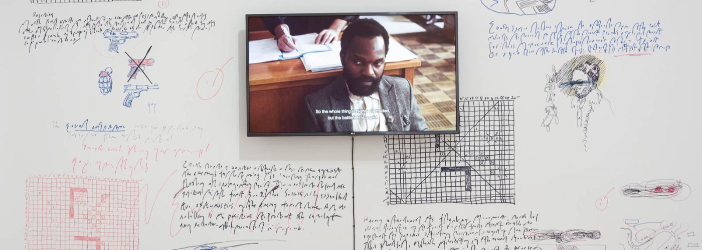 A television screen showing a close up of a black man looking towards the camera. The television screen is attached to a white wall which is covered in hand-drawn doodles and sketches in red, blue and black of scenes from comic strips.