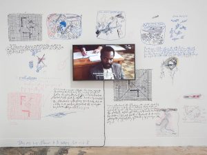 A television screen showing a close up of a black man looking towards the camera. The television screen is attached to a white wall which is covered in hand-drawn doodles and sketches in red, blue and black of scenes from comic strips.