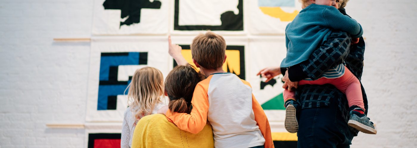 Two adults and three children facing away from the camera looking at a series of colourful fabric wall hangings above them. One adult wearing a yellow top stands next to two children and is pointing up at the hangings. Next to her another adult is holding a young child who is looking back towards the camera.