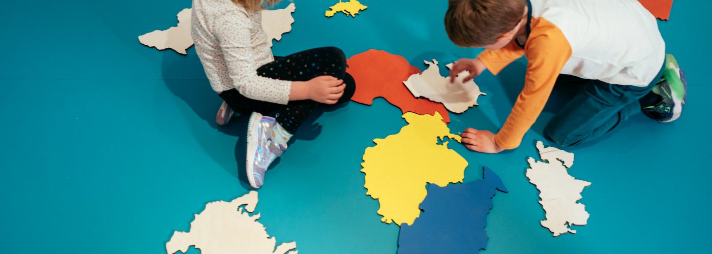 Two children on a bright blue floor playing with brightly coloured cut-outs of a map, viewed from above.
