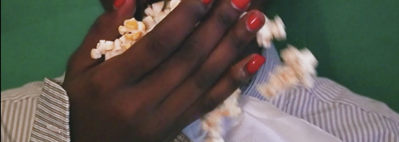 Close up shot of a Black woman putting a handful of popcorn into her mouth. Her hand covers most of her mouth and the popcorn is also falling out of her hand. She has bright red painted nails and is wearing a brown and white striped shirt. She has a white napkin tucked into her collar. She is standing against a dark green background.