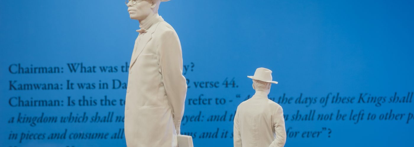 A sculpture of two men, one much larger than the other, both wearing hats, facing away from one another, standing on a plinth. Behind on a bright blue wall is black text, obscured by the statue, relaying a dialogue between Chairman and Kamwana.